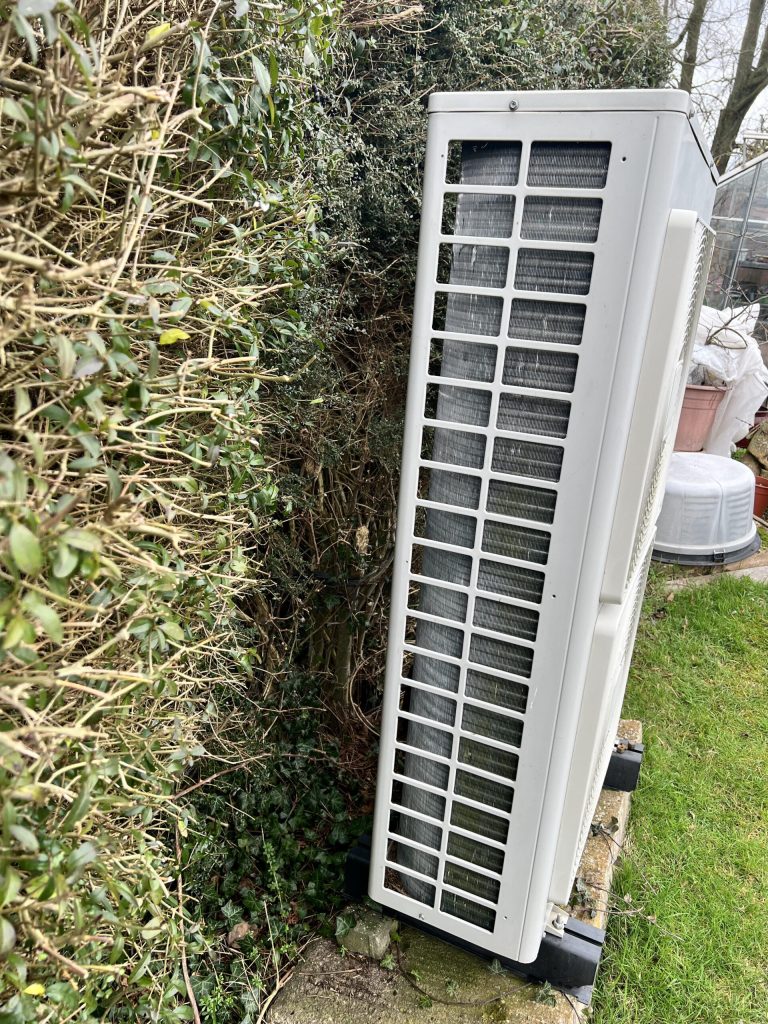 Side view of an air source heat pump beside a hedge