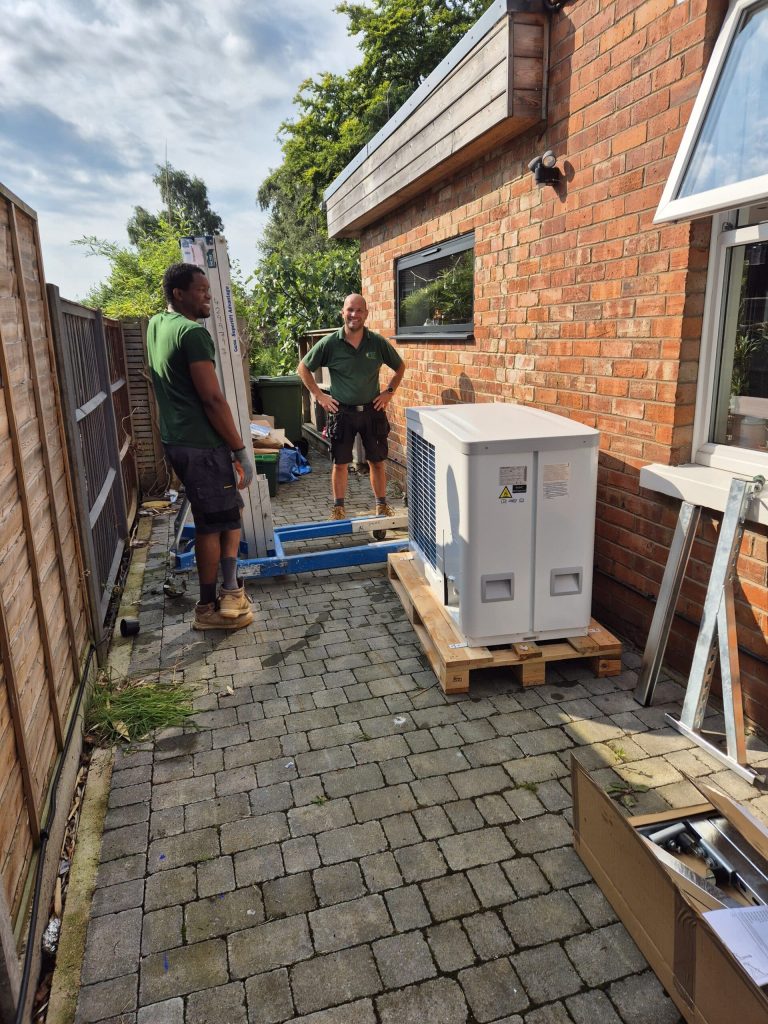 Installers delivering an air source heat pump beside a brick house